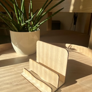 May include: A light-coloured wooden display stand featuring a tiered design with three distinct slots for holding items. The stand is constructed from smooth, flat plywood panels with rounded top corners, showcasing a natural wood grain texture.
