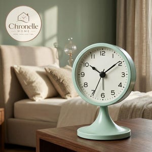 May include: A mint green analog table clock with a round face, white dial, and black numerals. The clock features black hour, minute, and second hands, and is supported by a matching mint green pedestal base. The design is simple and functional.