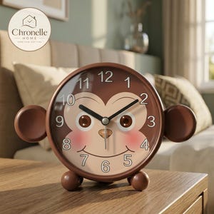 May include: A round brown analog alarm clock featuring a monkey face design on the dial with black hour and minute hands. The clock has two circular ears on the sides, two small round feet, and black numbers from 1 to 12.
