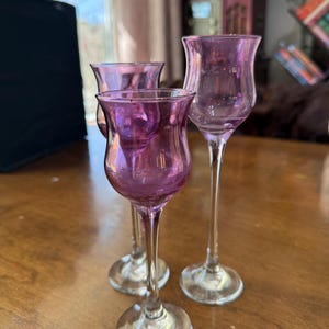 May include: Three purple glass cordial glasses with clear stems and circular bases. Each glass features a tulip-shaped bowl with a slightly flared rim. The glasses are arranged in a cluster, showcasing the translucent purple hue of the glass.