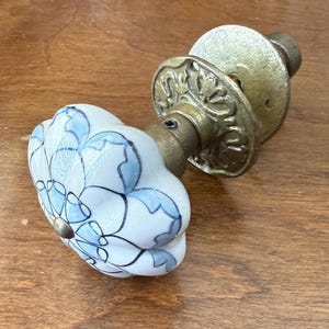 May include: A door knob featuring a white ceramic handle with a scalloped edge and a blue floral pattern. The handle is attached to an aged brass base plate with an intricate embossed design and a central metal screw fastener.