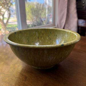 May include: A round, deep bowl with a smooth, glossy finish in a mottled olive green color. The surface features a speckled pattern with small flecks of darker green, yellow, and white throughout the material, creating a textured appearance.