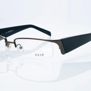 May include: Semi-rimless eyeglasses with a dark metallic brown frame and wide black temples. The right lens features the brand name exté printed in black. The bridge and nose pads are metallic brown, and the temples display the exté logo.