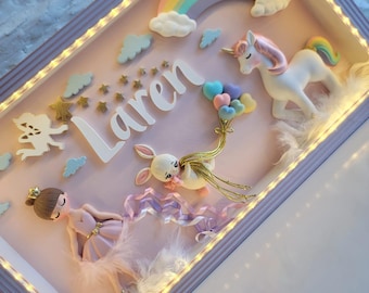 Puede incluir: Un marco de caja de sombra rectangular con luces LED internas que contiene una escena en 3D. Incluye el nombre Laren en letras blancas, un unicornio blanco, una bailarina con vestido rosa, un conejo con globos de corazón y un arcoíris.