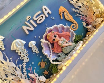 Puede incluir: Una caja de sombra con el nombre NISA en letras blancas, una figura de sirena en una concha, un caballito de mar, medusas, tortugas marinas y rayas. La escena incluye coral blanco y rosa, perlas e iluminación LED integrada.