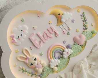 Puede incluir: Un aplique de pared con forma de nube que muestra el nombre Elay en letras rosas. El diseño incluye una luna creciente amarilla, una nube blanca con un lazo rosa, un arcoíris pastel, un conejo pequeño con un corazón y colinas verdes con estrellas.