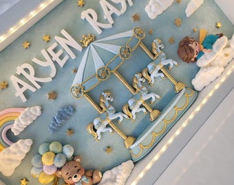 Puede incluir: Un marco tipo caja con luces LED que muestra el nombre ARYEN RAYAN en letras blancas. La escena incluye un carrusel con caballos blancos y dorados, un oso de peluche marrón con globos pastel, un arcoíris, nubes y una figura durmiendo.