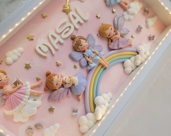 Puede incluir: Un marco tipo caja blanca con luces LED integradas que contiene un fondo rosa, el nombre MASAL en letras blancas, cuatro figuras de hadas, un arcoíris pastel, nubes blancas, estrellas doradas y pequeños detalles de mariposas.