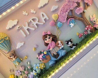 Puede incluir: Una caja decorativa con el nombre IKRA en letras blancas, una figura en un scooter azul con un cachorro, un globo aerostático con un oso, un árbol rosa, un gato blanco y negro y flores coloridas sobre una base verde con luces LED.