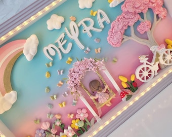 Puede incluir: Una caja decorativa con el nombre MEYRA en letras blancas, un arcoíris, un sol, nubes, un árbol de flores rosas, una bicicleta y una figura en un columpio. La escena incluye flores coloridas, mariposas y una valla blanca, todo iluminado por luces LED integradas.