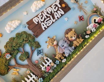 Puede incluir: Una caja de sombra con un letrero de madera que dice MEHMET ASLAN, rodeado de figuras de arcilla de un león, un elefante y una jirafa. La escena incluye un árbol, una valla blanca, nubes, globos y luces LED integradas.