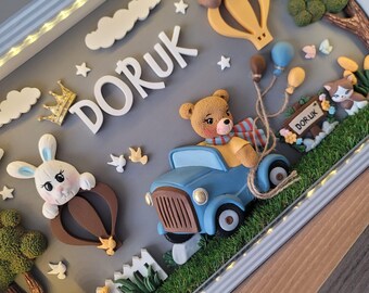 Puede incluir: Un marco tipo caja con una escena en 3D que muestra un coche azul conducido por un oso de peluche, un conejo blanco en un globo aerostático marrón, un oso en un globo amarillo, árboles, nubes, estrellas, una corona dorada y el nombre DORUK.