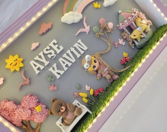 Puede incluir: Un marco tipo caja con borde morado claro y luces LED internas. La exhibición presenta el nombre AYŞE KAVIN en letras blancas, un arcoíris, un carrusel, un oso de peluche en un banco blanco, un árbol y un personaje con globos.
