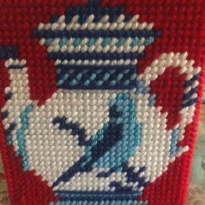 Pode incluir: Uma base de chá vermelha em ponto de cruz com um design detalhado de bule em branco, azul e azul claro. A base é feita de um material texturizado e costurado, mostrando uma estética clássica e artesanal.