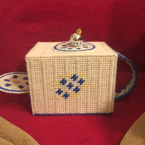 Op de afbeelding: Een witte gehaakte theepot met blauwe en gele details. De theepot heeft een zilveren ornament bovenop en een handvat.
