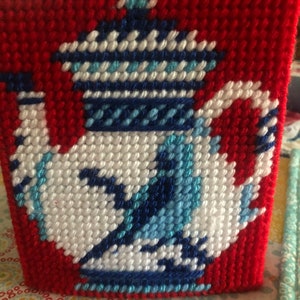 May include: A red canvas with a white and blue teapot design. The teapot has a blue and white patterned lid and a blue and white patterned handle.