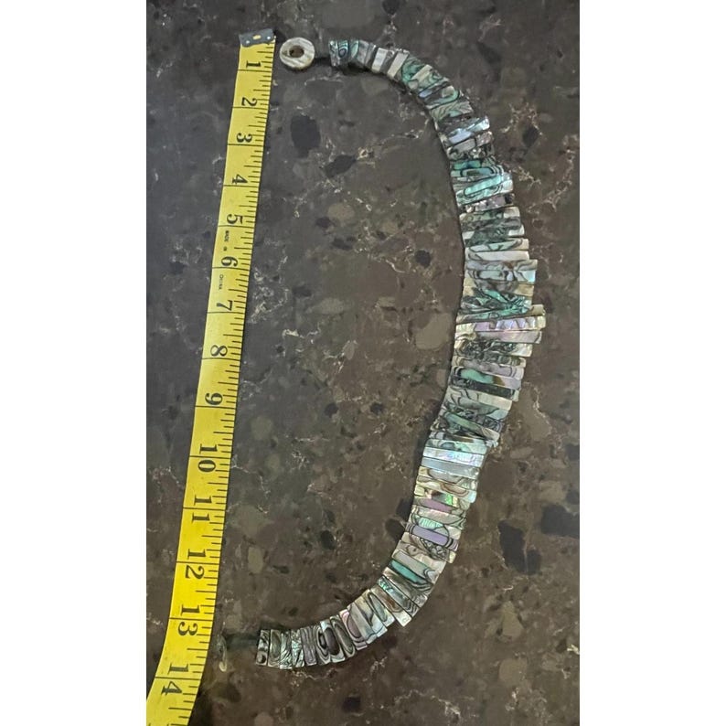 Pode incluir: Colar de concha de abalone com pe&ccedil;as retangulares iridescentes azuis, verdes e prateadas. O colar tem aproximadamente 33 cm de comprimento. Uma fita m&eacute;trica amarela est&aacute; ao lado do colar.