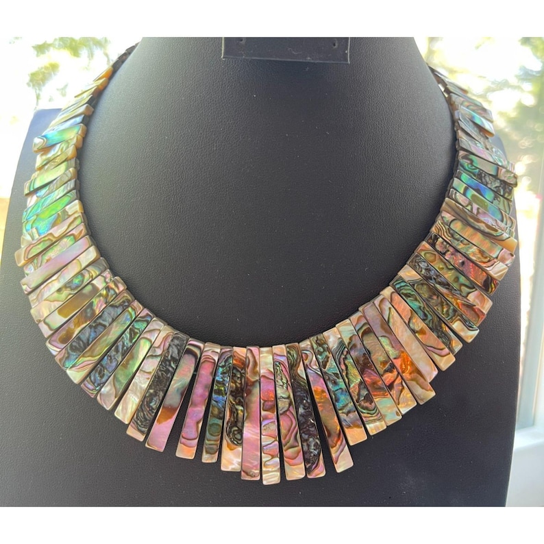 Pode incluir: Um colar marcante com segmentos retangulares iridescentes em tons de verde, roxo e dourado. O colar &eacute; exibido em um manequim preto, destacando seu design &uacute;nico. As pe&ccedil;as de concha s&atilde;o dispostas em um padr&atilde;o graduado.