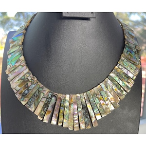 Pode incluir: Um colar marcante com pe&ccedil;as retangulares iridescentes de concha. O colar &eacute; exibido em um manequim preto. As pe&ccedil;as de concha exibem cores naturais, incluindo verdes, azuis e dourados. O design &eacute; um estilo gargantilha.