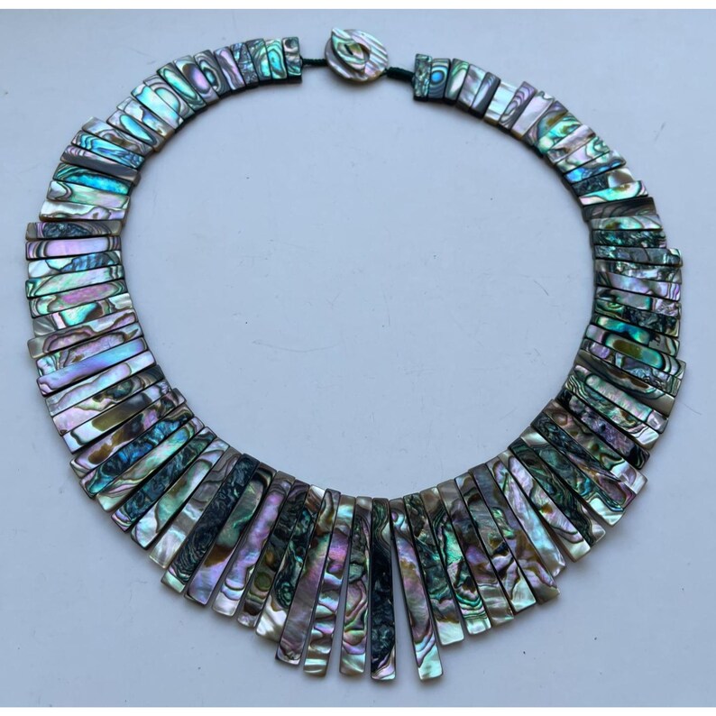 Pode incluir: Colar de concha de abalone iridescente com contas retangulares em tons de azul, verde e roxo. O colar tem um cord&atilde;o preto e um fecho de bot&atilde;o. As contas s&atilde;o dispostas em um design graduado.