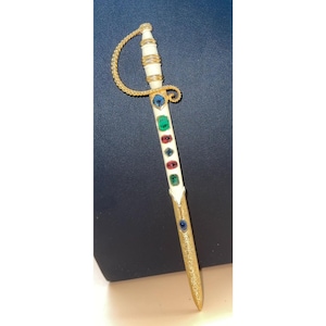 May include: A decorative letter opener in the shape of a miniature sword. The handle is white with a gold-colored hilt and guard. The blade is gold-colored and adorned with colorful gemstones, including blue, green, and red.