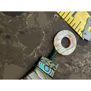 Pode incluir: Uma vista de perto de uma pulseira feita &agrave; m&atilde;o com contas de concha de abalone iridescentes e um pingente circular a condizer. O pingente e as contas t&ecirc;m um brilho perolado com toques de rosa e verde. Uma fita m&eacute;trica amarela est&aacute; no fundo.