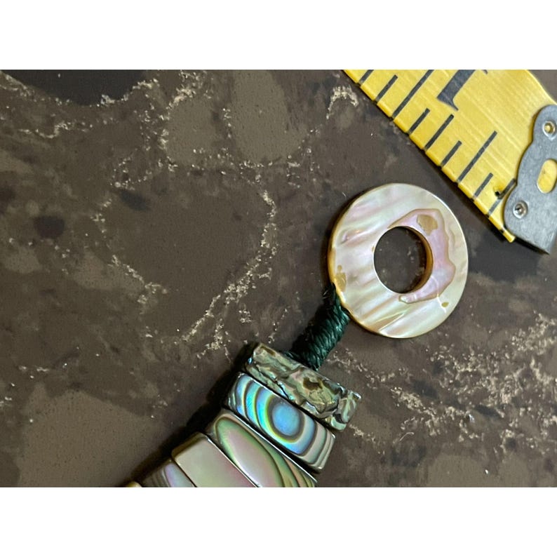 Pode incluir: Uma vista de perto de uma pulseira feita &agrave; m&atilde;o com contas de concha de abalone iridescentes e um pingente circular a condizer. O pingente e as contas t&ecirc;m um brilho perolado com toques de rosa e verde. Uma fita m&eacute;trica amarela est&aacute; no fundo.