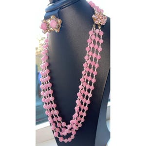 May include: A pink beaded necklace with matching earrings. The necklace has multiple strands of pink beads with silver spacers. The earrings and necklace clasp feature a flower design with pink accents. The jewelry is displayed on a black stand.