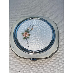 May include: A square silver-toned compact mirror with rounded corners. The center features a circular white enamel inlay with a textured guilloche pattern and a small painted floral motif consisting of orange flowers and green leaves.