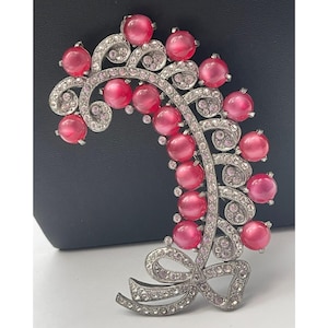 May include: A silver-toned brooch featuring a curved, leafy branch design adorned with numerous round, translucent pink cabochon beads and small, clear pavé-set rhinestones. The base of the branch is finished with a decorative ribbon bow.