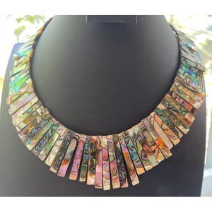 Pode incluir: Um colar marcante com pe&ccedil;as retangulares iridescentes de concha em tons de verde, rosa e dourado. O colar &eacute; exibido em um manequim preto, mostrando seu design &uacute;nico e atraente. As pe&ccedil;as de concha s&atilde;o dispostas em um padr&atilde;o graduado.