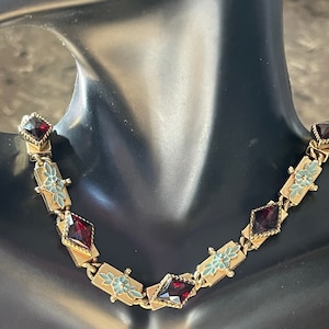 May include: A gold necklace with red and blue enamel accents. The necklace features a repeating pattern of small flowers and red gemstone-like pieces.