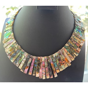 Pode incluir: Um colar marcante com pe&ccedil;as retangulares iridescentes de concha em tons de rosa, verde e dourado. O colar &eacute; exibido em um manequim preto, destacando as cores vibrantes e o design &uacute;nico. As pe&ccedil;as de concha s&atilde;o dispostas em um padr&atilde;o graduado.