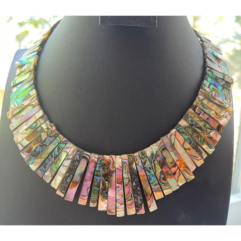 Pode incluir: Um colar marcante com pe&ccedil;as retangulares iridescentes de concha em tons de rosa, verde e dourado. O colar &eacute; exibido em um manequim preto, destacando as cores vibrantes e o design &uacute;nico. As pe&ccedil;as de concha s&atilde;o dispostas em um padr&atilde;o graduado.