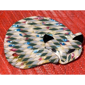 May include: A decorative fox brooch in a sleeping pose. The brooch features a geometric pattern in shades of gray, green, white, and pink, with gold accents. The fox's ears are black. The brooch is set against a red textured background.