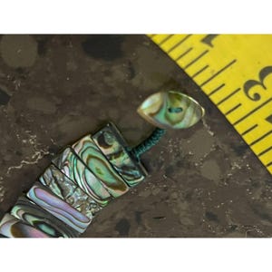 Pode incluir: Pulseira de concha de abalone com cores iridescentes de verde, azul e roxo. A pulseira apresenta pe&ccedil;as retangulares de concha unidas por um cord&atilde;o verde. O fecho &eacute; um bot&atilde;o oval com um design de concha de abalone correspondente.