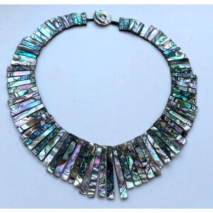 Pode incluir: Colar de concha de abalone com cores iridescentes de azul, verde e roxo. O colar apresenta pe&ccedil;as retangulares de concha, criando um design em forma de leque. O fecho &eacute; uma pequena pe&ccedil;a redonda de concha.