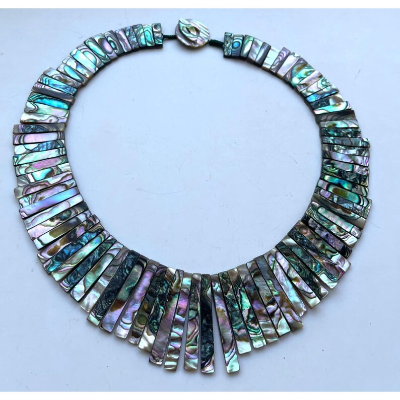 Pode incluir: Colar de concha de abalone com cores iridescentes de azul, verde e roxo. O colar apresenta pe&ccedil;as retangulares de concha, criando um design em forma de leque. O fecho &eacute; uma pequena pe&ccedil;a redonda de concha.