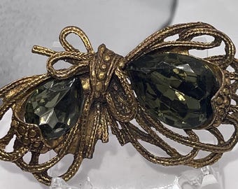 May include: A gold-toned brooch with a bow design and two large, dark green faceted glass stones.