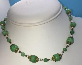 Puede incluir: Un collar vintage con cuentas verdes moteadas, intercaladas con cuentas blancas y detalles dorados. El collar se exhibe sobre un busto de maniquí blanco, resaltando su diseño y colores.