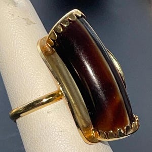 May include: A gold ring with a large, dark brown, oval-shaped stone set in a gold setting. The stone has a textured surface and is surrounded by a gold border.