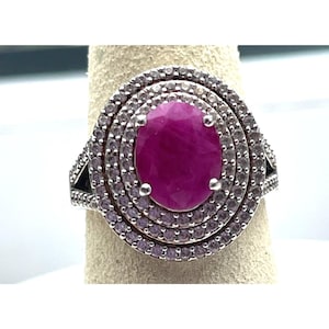 May include: A silver-toned ring featuring a large, oval-cut, deep pink center stone. The center stone is surrounded by two concentric halos of small, clear, round-cut stones. The band is split and decorated with additional small, clear stones.