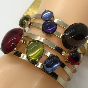 May include: A set of three gold bangle bracelets with colorful glass cabochon stones. The stones are in shades of red, green, blue, and purple.