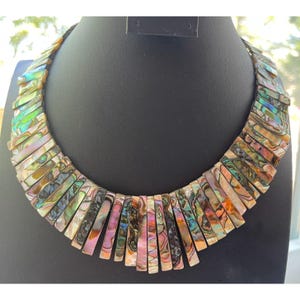 Pode incluir: Um colar marcante composto por segmentos retangulares iridescentes de concha de abalone. As pe&ccedil;as de concha exibem uma gama de cores, incluindo verde, rosa e dourado, dispostas em um padr&atilde;o graduado. O colar &eacute; exibido em um manequim preto.