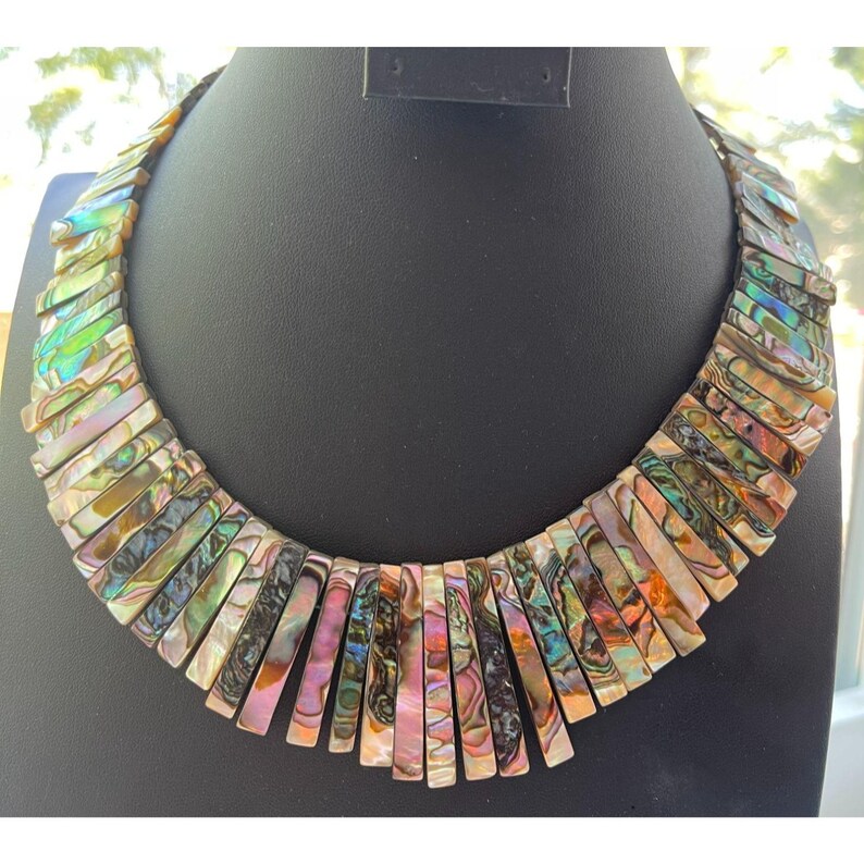 Pode incluir: Um colar marcante composto por segmentos retangulares iridescentes de concha de abalone. As pe&ccedil;as de concha exibem uma gama de cores, incluindo verde, rosa e dourado, dispostas em um padr&atilde;o graduado. O colar &eacute; exibido em um manequim preto.
