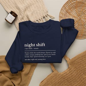 May include: A navy blue crewneck sweatshirt featuring white text that defines the term night shift. The text includes a phonetic pronunciation, a noun label, and a humorous description of working while the world sleeps, plus see also references.