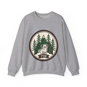 Può includere: Una felpa girocollo grigio melange con una grafica circolare sul petto. Il disegno raffigura un piccolo cane bianco seduto davanti a pini verdi e cime montuose, con un banner marrone scuro nella parte inferiore contenente il testo PNW.