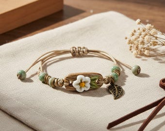 May include: A beige cord bracelet featuring a central wooden-textured bead with a white flower and green leaves, accented by light green beads, small metallic rings, and a dangling bronze-toned leaf charm with an adjustable sliding knot closure.