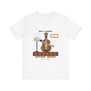 Könnte beinhalten: Ein weißes T-Shirt mit einer Pixel-Art-Grafik eines braunen Cellos mit Bogen, einem Notenständer mit Noten und dem Text BOW CLEARED, ENCORE BONUS und FIRST CHAIR. Das Design enthält eine braune Plattform und kleine dekorative Elemente.