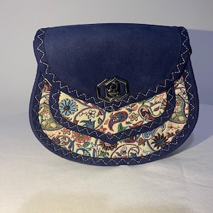 Può includere: Una piccola borsa in camoscio blu navy con chiusura a patta dotata di una chiusura a girello esagonale in metallo. Il pannello frontale presenta un tessuto color crema con un motivo floreale e paisley multicolore, accentuato da cuciture a zigzag.